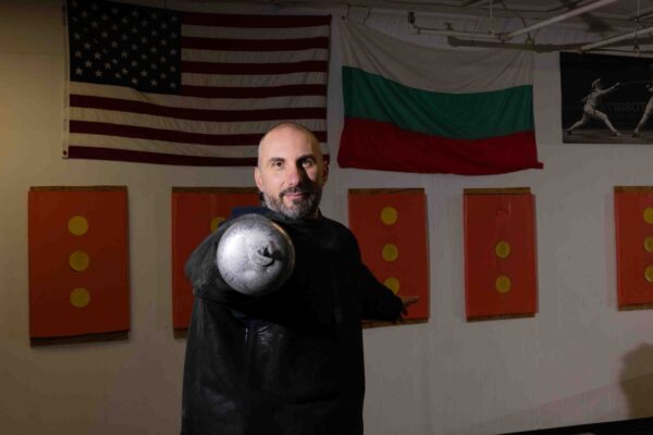 A fencer dressed in black stands in a fencing studio with his sword pointed towards the camera.