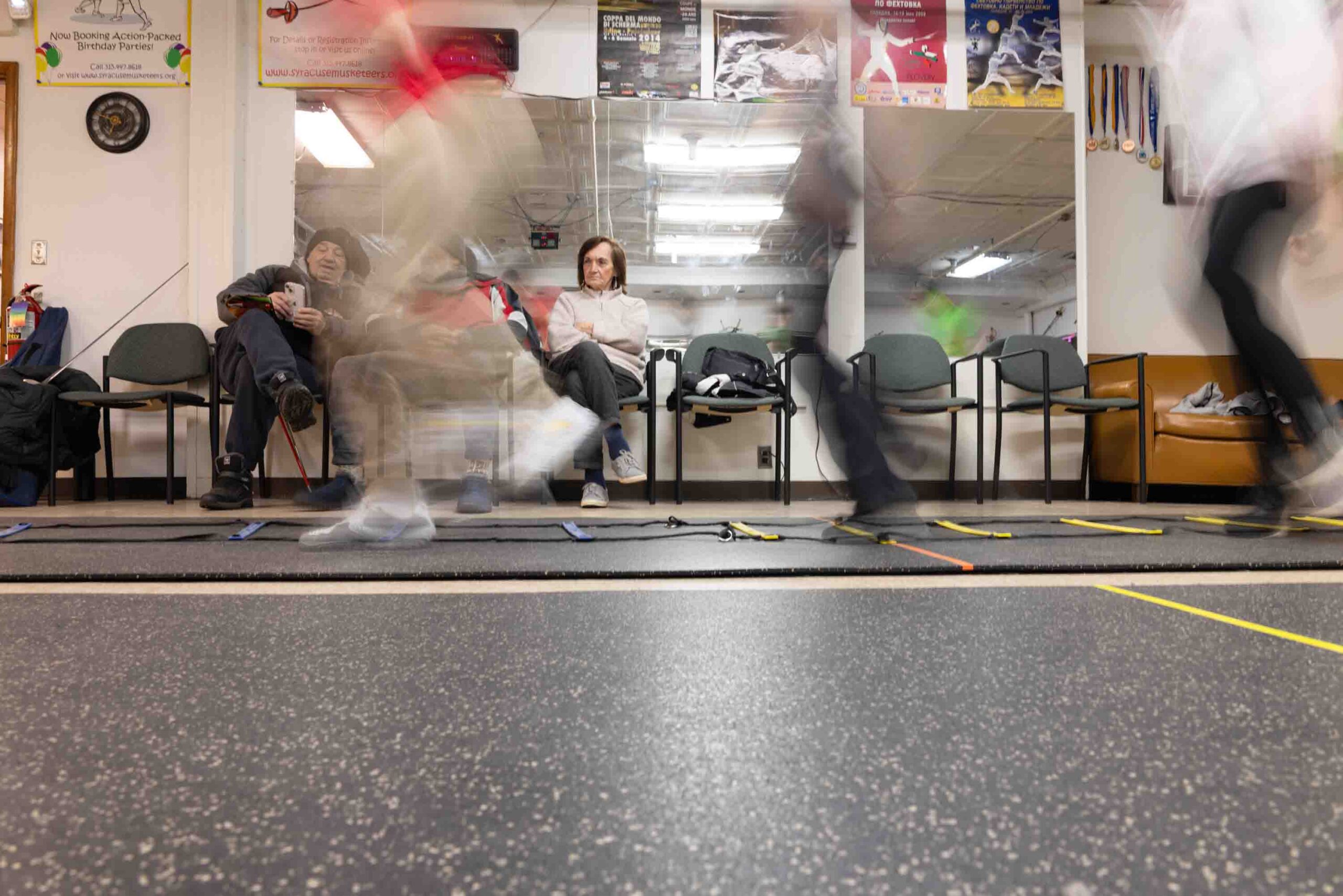 In a fencing studio, parents look on as the students practice movement.