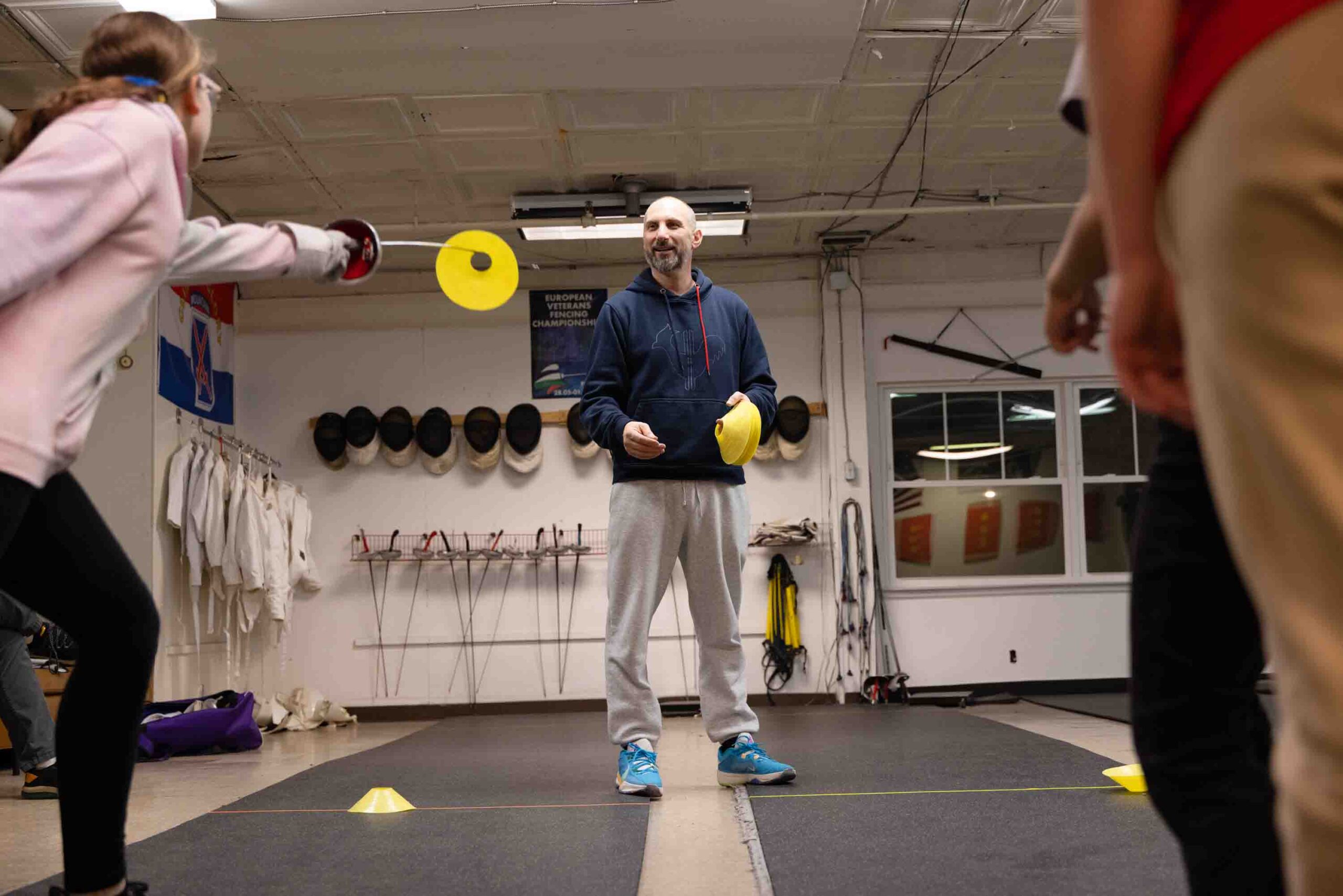 A coach stands in a fencing studio and throws yellow disks in the air for the students to catch on their swords.