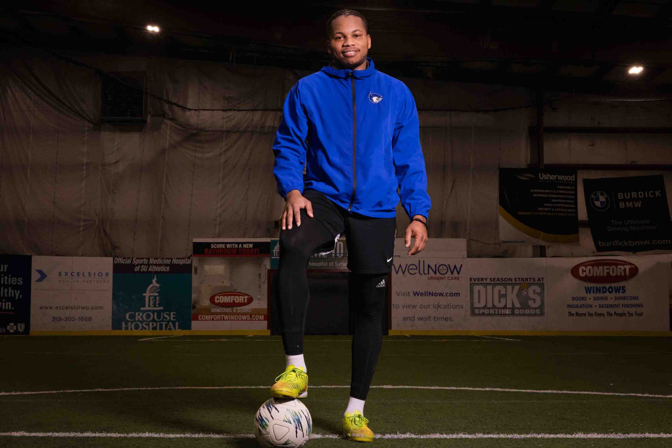 In this photo Jhonny Zamor stands in a blue zip up on an indoor soccer field with his right footing resting on a soccer ball.