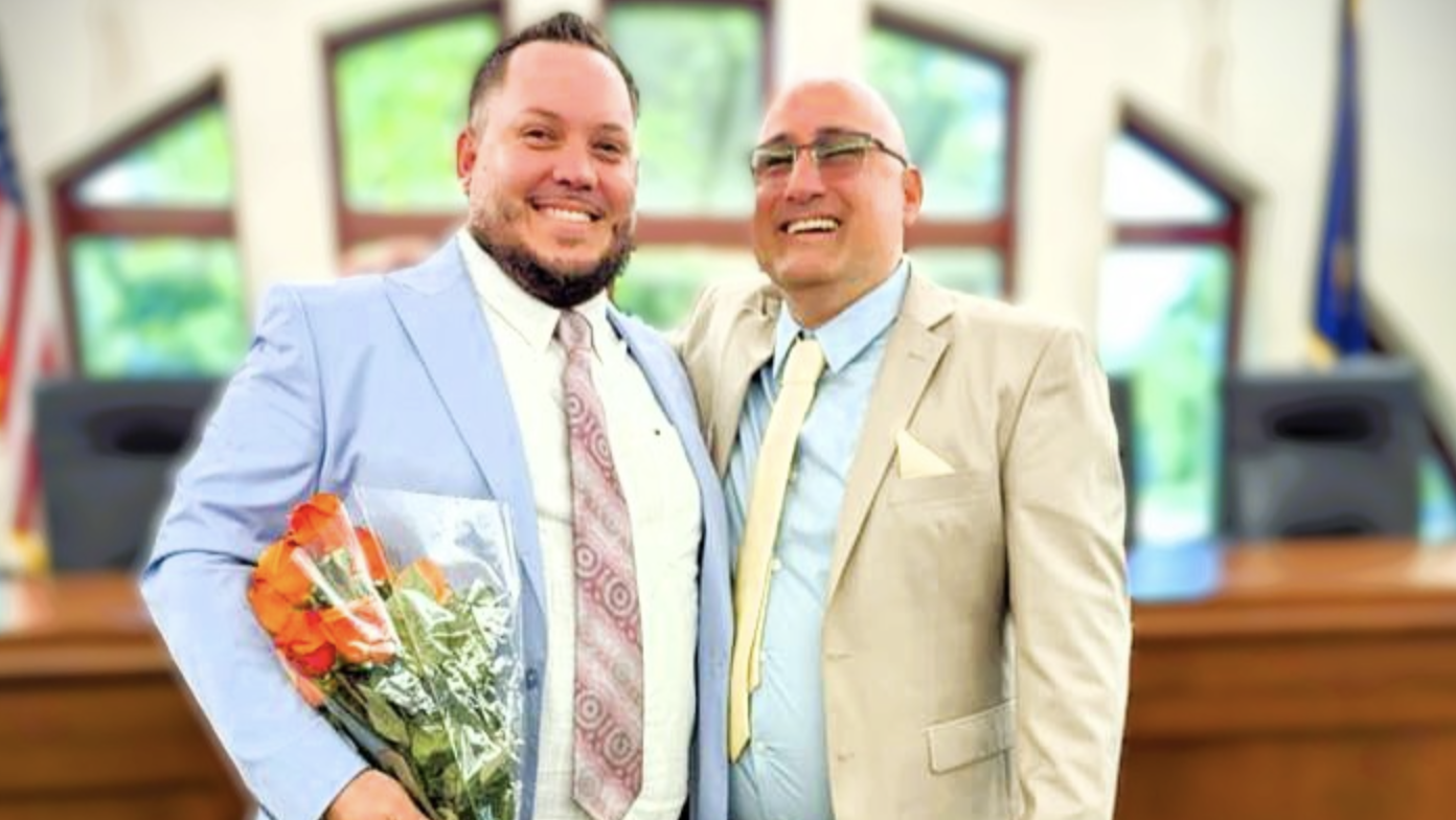 Alex Gonzalez and Yan Vasquez stand in suits in a courtroom smiling with their arms around each other.