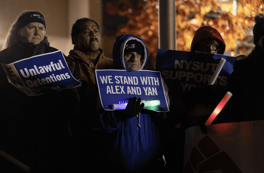 At a night protest, protesters stand with blue signs that read "We stand with Alex and Yan."