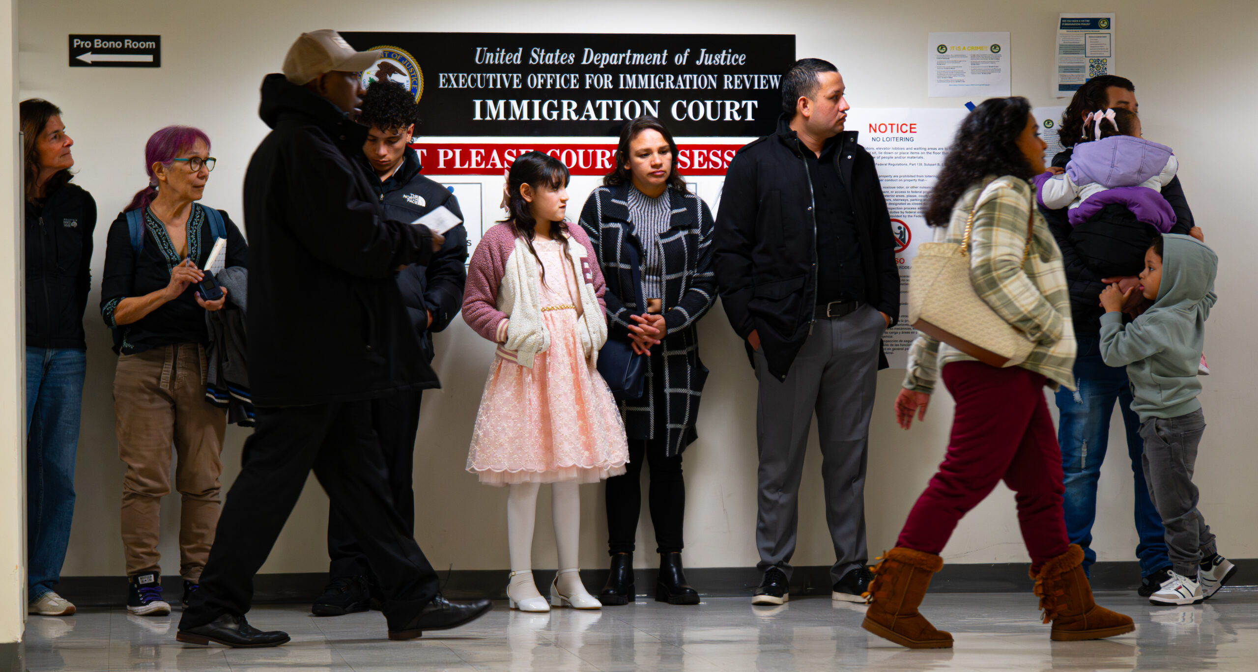 Under a sign that reads "United States Department of Justice Immigration Court" a number of people wait in line to head inside.